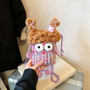 Könnte beinhalten: Eine skurrile, handgefertigte, mehrfarbige Stricktasche mit großen Augen mit weißem Rand und einem flauschigen braunen Oberteil. Die Tasche hat lila hängende Akzente und eine kleine Blume. Die Tasche wird in der Hand einer Person gehalten.