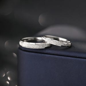 Puede incluir: Dos anillos de plata con un diseño de banda texturizada. Los anillos se muestran sobre una superficie azul oscuro, con un fondo borroso. Los anillos tienen un aspecto brillante y metálico, lo que sugiere que están hechos de un metal precioso.