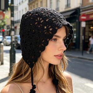 Op de afbeelding: Een zwarte gehaakte hoed met een bloemenpatroon. De hoed heeft lange banden met bloemvormige accenten. De hoed wordt gedragen door een persoon met lang blond haar.