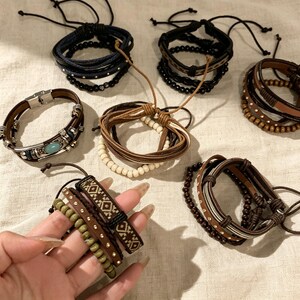 Könnte beinhalten: Eine Sammlung handgefertigter Armbänder in verschiedenen Stilen. Die Armbänder bestehen aus Leder, Perlen und geflochtenem Kordel in Braun-, Schwarz- und Blautönen. Einige Armbänder haben dekorative Metallakzente und verstellbare Verschlüsse.
