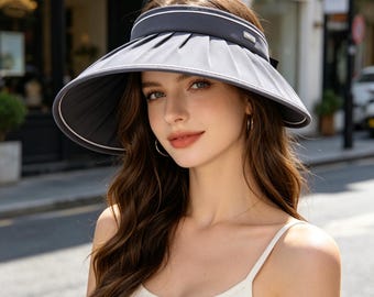 May include: A grey sun visor with white trim and a dark grey band. The visor is pleated and has a wide brim. The hat is designed to protect the face from the sun.