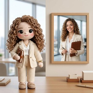 Puede incluir: Mu&ntilde;eca de ganchillo con un traje de negocios color crema, sosteniendo un cuaderno marr&oacute;n. La mu&ntilde;eca tiene cabello casta&ntilde;o rizado y una cara sonriente. Una foto enmarcada de una mujer con un traje similar est&aacute; en la pared. La mu&ntilde;eca est&aacute; sobre una superficie de madera.