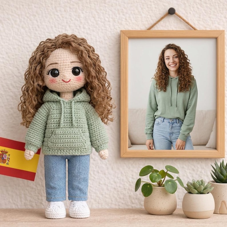 Puede incluir: Una mu&ntilde;eca de ganchillo hecha a mano con cabello rizado casta&ntilde;o, vestida con una sudadera con capucha verde, vaqueros azules y zapatillas blancas. La mu&ntilde;eca est&aacute; de pie junto a una foto enmarcada de una mujer con un atuendo similar. Se ve una bandera espa&ntilde;ola.