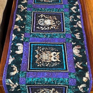 Puede incluir: Camino de mesa acolchado con diseños de búhos. El camino de mesa tiene un fondo verde oscuro con detalles morados y negros. Los paneles centrales muestran ilustraciones detalladas de búhos, mientras que los bordes muestran búhos volando y lunas crecientes.