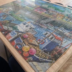Peut inclure: Puzzle terminé représentant un port avec des bateaux et des bâtiments. Le puzzle est encadré dans un cadre en bois clair. Les pièces du puzzle sont emboîtées, révélant une image détaillée et colorée du port.