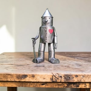 Puede incluir: Una figura metálica de color plateado que representa un robot con un sombrero cónico, un torso cilíndrico y extremidades articuladas. La figura presenta un pequeño emblema de corazón rojo en el lado izquierdo del pecho y sostiene un hacha.