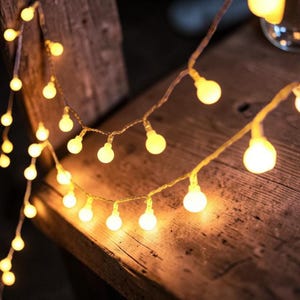 Puede incluir: Una cadena de pequeñas luces LED redondas de color blanco cálido. Las luces están ensartadas en un cable fino y transparente y están dispuestas sobre una superficie de madera rústica. Las luces brillan, creando un ambiente acogedor e invitador.