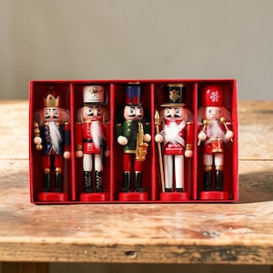 Puede incluir: Un conjunto de cinco cascanueces de madera en una caja roja. Cada cascanueces está pintado con detalles intrincados, incluyendo uniformes, sombreros y rasgos faciales. Los cascanueces son de varios colores, incluyendo rojo, azul, verde y dorado.