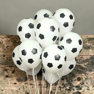 May include: A cluster of white latex balloons, each printed with a black pentagon pattern resembling a soccer ball. The balloons are round, inflated, and feature thin white ribbons attached to the base of each balloon for holding or tying.