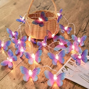 May include: A string of decorative butterfly lights with pink and blue wings, resting on a wooden surface. The butterflies are arranged around a small woven basket. The lights are on, creating a warm glow.