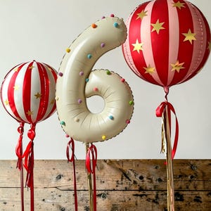 May include: A cream-colored number six balloon decorated with small multicolored pom-poms, flanked by two round balloons featuring red and light pink vertical stripes with gold star accents, each attached to red ribbons and gold sticks.