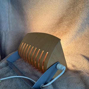 Peut inclure: Lampe de bureau en bois avec une base en métal bleu clair et un cordon blanc. La lampe a un abat-jour incurvé en bois avec des fentes verticales qui émettent une lueur chaude. La lampe est conçue pour être posée sur un bureau ou une table.