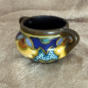 Puede incluir: Un cuenco de cerámica pequeño y redondo con dos asas. El cuenco presenta un interior azul oscuro y está decorado con un colorido estampado floral en tonos azules, amarillos y naranjas. El borde y las asas son de color marrón oscuro.