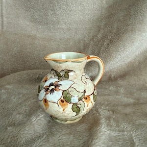 May include: A small, round ceramic pitcher with a handle and spout. The pitcher is painted with a floral design featuring white flowers with brown centers and green leaves. The background is a beige color.