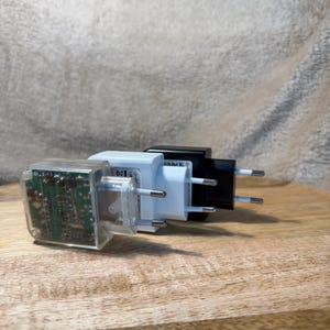 Könnte beinhalten: Drei Wandnetzadapter in einer Reihe, bestehend aus einem transparenten Kunststoffgehäuse mit sichtbarer grüner Platine, einem Gehäuse aus weißem Kunststoff und einem Gehäuse aus schwarzem Kunststoff, jeweils mit zwei Metallstiften für Steckdosen.