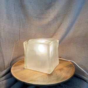 May include: A square, translucent glass lamp shaped like a large ice cube. The lamp emits a soft, warm white light from within and features an irregular, textured surface. A thin white power cord extends from the side of the glass cube base.