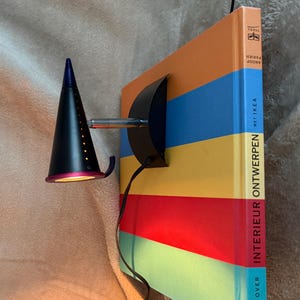 Könnte beinhalten: Eine schwarze, kegelförmige Wandleuchte mit blauer Spitze und pinkfarbenem Rand, die eine Reihe kleiner Lichtperforationen aufweist. Sie ist an einem Buch mit dem Titel INTERIEUR ONTWERPEN MET IKEA befestigt, das horizontale Streifen in Orange, Blau, Gelb, Rot und Türkis zeigt.