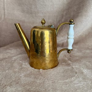 Op de afbeelding: Een vintage messing theepot met een witte handgreep. De theepot heeft een lange tuit en een deksel met een decoratieve knop. Het messing heeft een gehamerde textuur, wat het een rustieke uitstraling geeft. De handgreep is wit met messing accenten.