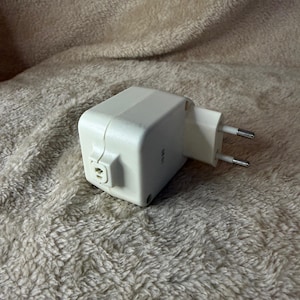 Peut inclure: Un adaptateur électrique blanc avec une prise à deux broches. L'adaptateur est rectangulaire avec des bords arrondis et un petit port sur un côté. Le fond est un tissu beige doux et texturé.