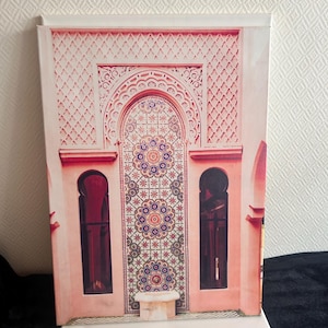 Peut inclure: Impression sur toile décorative représentant une arche de style marocain ornée de mosaïques roses, rouges et bleues. L'impression présente deux fenêtres arquées et une bordure supérieure détaillée.