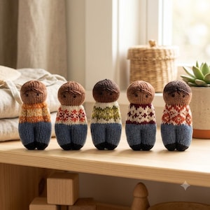 Könnte beinhalten: Fünf kleine, handgestrickte Puppen stehen in einer Reihe. Jede Puppe hat einen runden Kopf mit gestickten Gesichtszügen, einen gemusterten Pullover in verschiedenen Farben und eine blaue Hose mit schwarzen Füßen. Die Puppen sind aus Garn gefertigt.