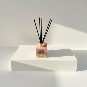 May include: A pink, cube-shaped essential oil diffuser with a gold-colored metal collar holding black reeds. The front of the diffuser has a gold tree design. The diffuser sits on a white, rectangular platform.