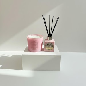 May include: A pink candle and reed diffuser set. The candle has a label with text, and the diffuser has a gold tree design. Black reeds are inserted into the diffuser. Both items are on a white block.