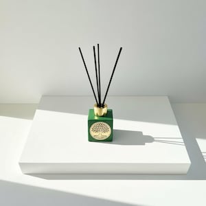 Puede incluir: Un difusor cuadrado verde con una tapa dorada y un emblema dorado del árbol de la vida. Las cañas negras se insertan en la botella. El difusor se encuentra sobre una superficie blanca.