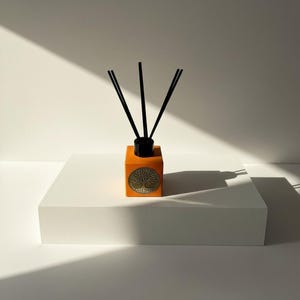 Könnte beinhalten: Ein orangefarbener, quadratischer Diffusor mit schwarzen Stäbchen. Die Flasche hat ein goldenes Lebensbaum-Emblem. Der Diffusor steht auf einer weißen, quadratischen Plattform vor einem weißen Hintergrund.