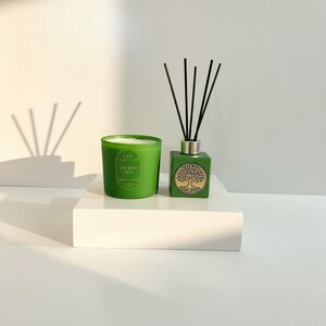 Könnte beinhalten: Ein grünes Kerzen- und Diffusor-Set. Die Kerze befindet sich in einem runden Glasbehälter mit dem Text "Sacred Oud". Der Diffusor ist quadratisch mit einem Baumdesign und schwarzen Stäbchen.