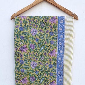 May include: A rectangular fabric textile featuring a light olive green background with a repeating floral pattern in shades of purple, blue, and green. The fabric has a blue floral border and a thin green and white striped edge on the right side.