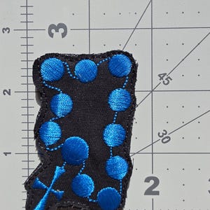 May include: Black patch embroidered with a blue rosary design. The rosary features blue beads and a cross at the bottom. The patch is approximately 2 inches wide and 3 inches tall.