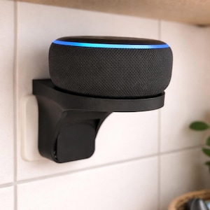 Könnte beinhalten: Ein schwarzer, kreisförmiger Smart-Lautsprecher mit einem leuchtend blauen Lichtring am oberen Rand, montiert auf einer schwarzen Kunststoff-Wandhalterung, die direkt in eine Steckdose an einer weißen Fliesenwand eingesteckt ist.