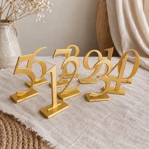 Könnte beinhalten: Eine Sammlung freistehender, goldfarbener Tischnummern mit eleganter, geschwungener Typografie. Jede Zahl ist auf einem rechteckigen Sockel montiert, was für Stabilität auf ebenen Flächen sorgt. Das Set enthält verschiedene Ziffern in Metallic-Optik.