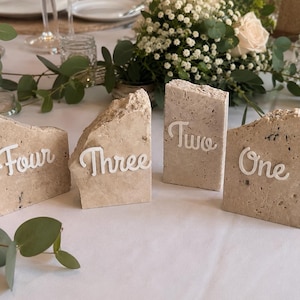 Puede incluir: Cuatro números de mesa rectangulares hechos de piedra travertino beige con bordes superiores rugosos e irregulares. Cada pieza presenta letras blancas en cursiva que deletrean las palabras One, Two, Three y Four, sobre una superficie blanca.