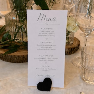 May include: A white rectangular menu card held by a black heart-shaped stand. The text reads Menü, Vorspeise, Carpaccio vom Rind, Hauptgang, Rinderfilet, Vegetarisch, Risotto, Dessert, Panna Cotta, and Guten Appetit. The card is printed in black ink.