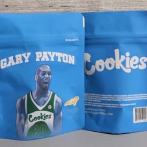 Pode incluir: Duas sacolas azuis, sel&aacute;veis, com o texto "GARY PAYTON" e "Cookies" em branco. Uma sacola apresenta um gr&aacute;fico de um jogador de basquete com uma camisa verde e branca. As sacolas t&ecirc;m um c&oacute;digo de barras e outro texto.