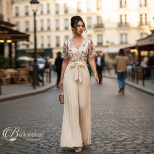 Könnte beinhalten: Cremefarbener Jumpsuit mit floral besticktem Oberteil und weiten, plissierten Beinen. Das kurzärmelige Oberteil hat einen V-Ausschnitt und einen Bindegürtel in der Taille. Das Outfit wird mit einer kleinen, gemusterten Handtasche ergänzt.