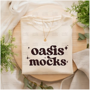 May include: A cream-colored t-shirt featuring the dark brown text oasis mocks in a retro font, accented by four-pointed star icons. A delicate gold-toned chain necklace with a small circular pendant rests on the neckline of the shirt.