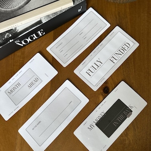 May include: Five white rectangular cards with text, including "MONTH AHEAD", "IN THE BANK", and "FULLY FUNDED". The cards are arranged on a wooden surface, with a book and decorative glass in the background.