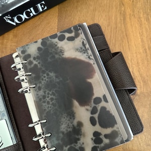 Könnte beinhalten: Ein brauner Lederplaner mit silbernen Binderingen und einem gemusterten Einband. Der Einband hat eine durchscheinende Auflage mit dunkelgrauen und braunen Flecken. Das Wort "VOGUE" ist auf einem Buch im Hintergrund zu sehen.