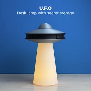 May include: A desk lamp shaped like a UFO, with a gray top and a white, cone-shaped base that emits a warm glow. The lamp has the text "U.F.O Desk lamp with secret storage" in white letters against a blue background.