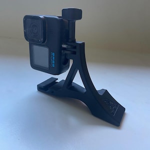 Puede incluir: Una cámara de acción GoPro negra montada sobre un soporte de plástico negro impreso en 3D con formas angulares. La cámara cuenta con una lente cuadrada, una pantalla frontal y el logotipo de GoPro impreso en azul en el lateral. El soporte tiene una base curva.