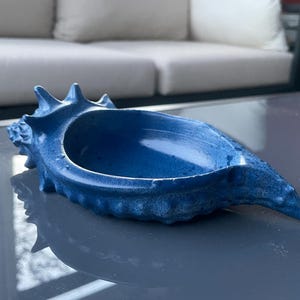 Puede incluir: Un cuenco decorativo azul con forma de concha. El cuenco tiene una superficie texturizada y un interior profundo y abierto. El color es un azul vibrante y sólido, y el cuenco descansa sobre una superficie reflectante. El diseño de concha añade un tema costero o náutico.
