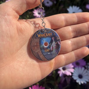 May include: A round keychain featuring a World of Warcraft DVD design. The keychain has a silver metal chain and ring. The DVD design includes the game's logo and artwork of a character. The background is a blur of purple and white flowers.