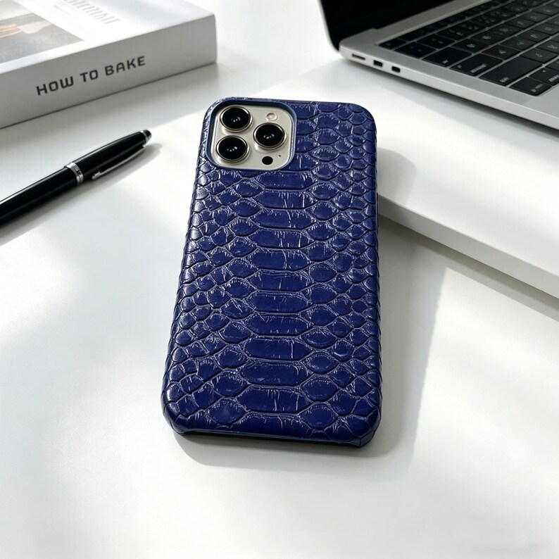 May include: A navy blue phone case with a textured, snake-skin pattern. The case has a camera cutout and is lying on a white surface. A black pen and a book titled "HOW TO BAKE" are also visible.