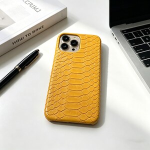 May include: Mustard yellow phone case with a textured snakeskin pattern. The case has a camera cutout for multiple lenses. A black pen and a book with the text "HOW TO BAKE" are also visible.