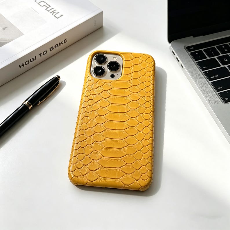May include: Mustard yellow phone case with a textured snakeskin pattern. The case has a camera cutout for multiple lenses. A black pen and a book with the text "HOW TO BAKE" are also visible.