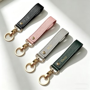 May include: Four personalized keychains in black, pink, gray, and green. Each keychain features a leather strap with gold hardware and a split ring. The black keychain has "A.C.E" printed on it, the gray keychain has "M.G", and the green keychain has "Daisy".