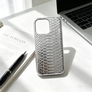 May include: Silver phone case with a snakeskin texture. The case has a cutout for the camera and buttons. The phone case is on a white surface next to a laptop and an open book with the text "THE LOVCED".